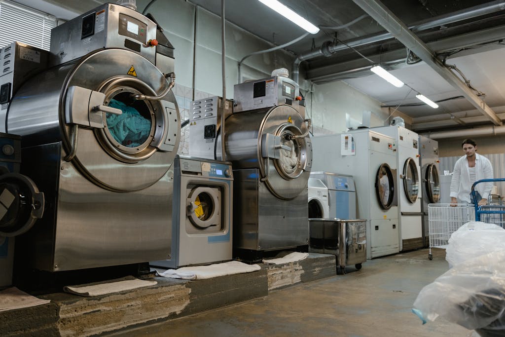 Industrial laundromat interior with large washing machines and a man pushing a trolley.